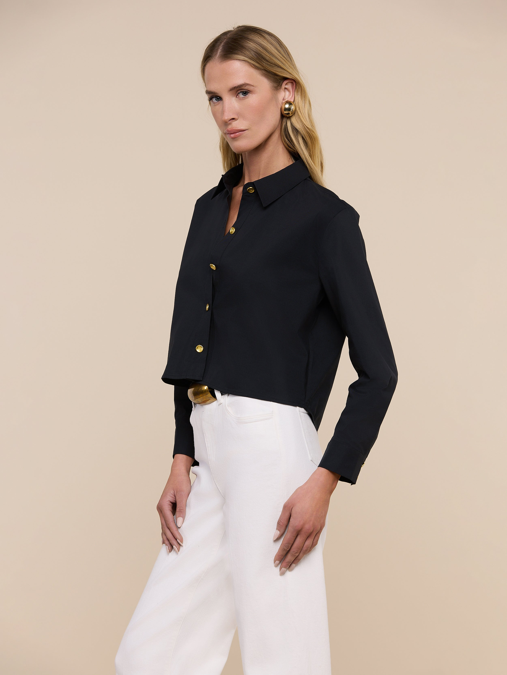 A woman with blonde hair wears the Cosette Shirt—a tailored black button-up—paired with blue jeans, standing against a plain light background and facing slightly to the side.