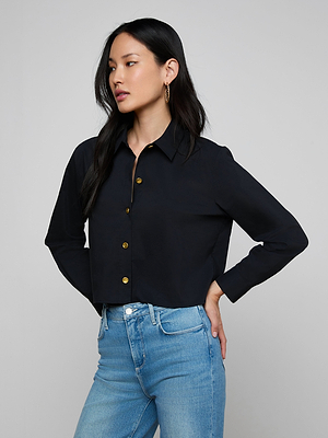 A woman with long black hair wears the Cosette Shirt, a tailored black button-up, paired with light blue jeans. She stands with one hand on her hip, looking to the side against a plain gray background.