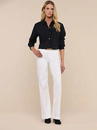 A woman with long blonde hair wears sunglasses, the Cosette Shirt—a tailored black button-up—blue jeans, and black heels as she stands against a plain white background.