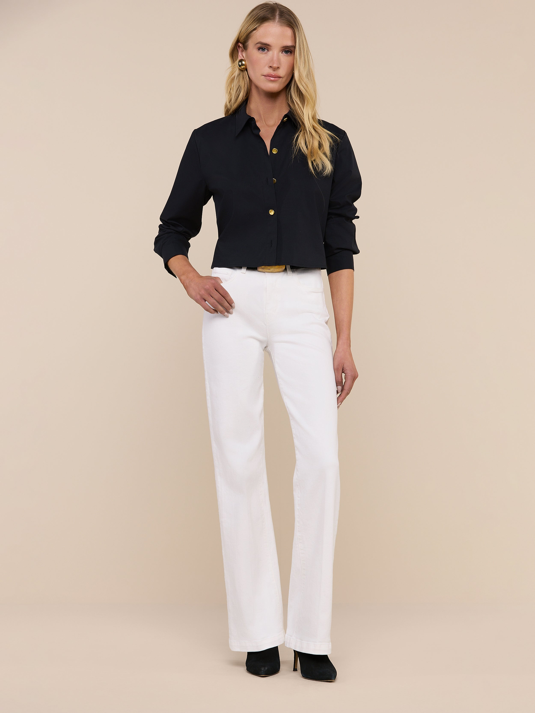 A woman with long blonde hair wears sunglasses, the Cosette Shirt—a tailored black button-up—blue jeans, and black heels as she stands against a plain white background.