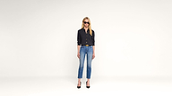 A woman wearing sunglasses, a black buttoned shirt, blue jeans, and black heels stands facing forward against a plain white background.