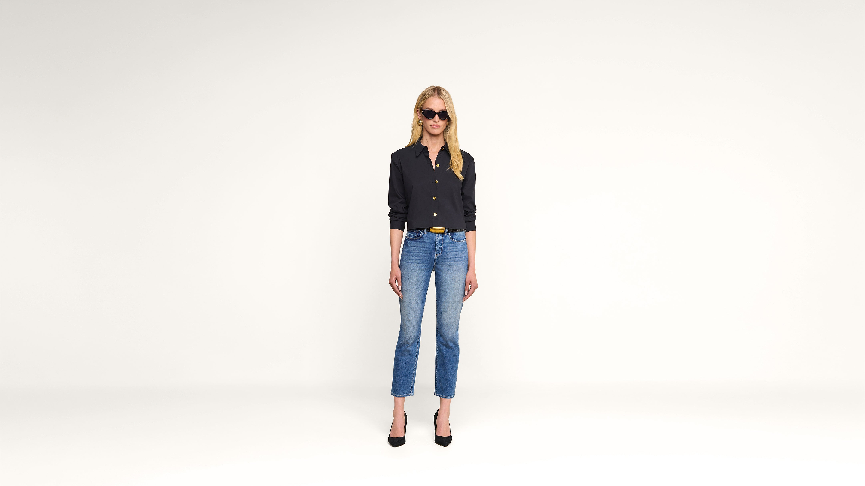 A woman wearing sunglasses, a black buttoned shirt, blue jeans, and black heels stands facing forward against a plain white background.