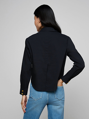 A woman with long dark hair wears the Cosette Shirt, a black tailored top with a cropped hem, paired with light blue jeans. She stands with her back to the camera, left hand on hip, against a plain light gray background.