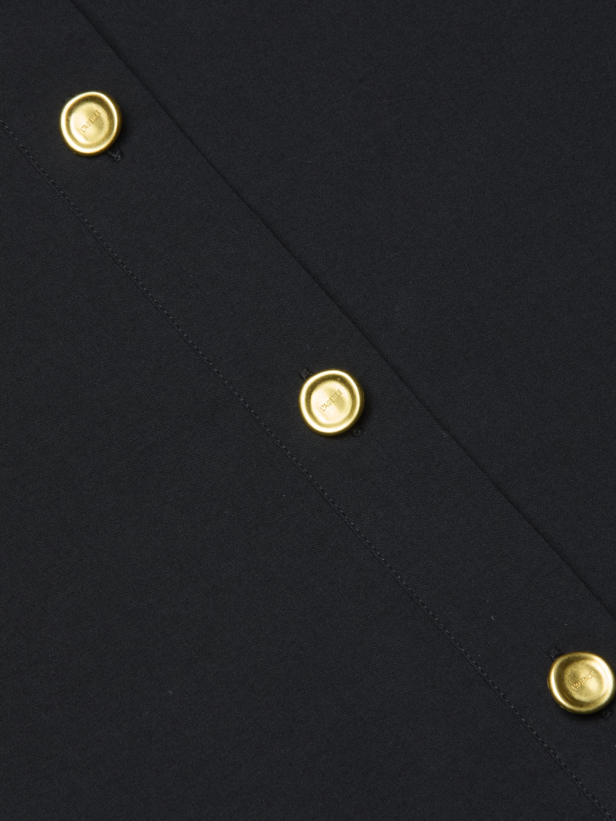 Close-up of the Cosette Shirt in black organic cotton fabric, featuring a row of shiny gold round buttons evenly spaced along a stitched seam.
