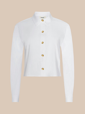The Cosette Shirt is a white poplin long-sleeve with a collared neckline, gold buttons, and a cropped high-low hem, shown on a plain beige background.