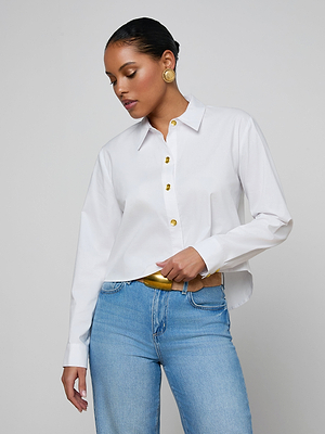 A woman wearing the Cosette Shirt—white organic cotton poplin with gold buttons—paired with blue jeans, gold earrings, and a gold belt stands against a plain light background, looking down with one hand in her pocket.