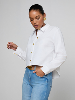 A woman models the Cosette Shirt, a white organic cotton poplin top, paired with blue jeans. With her hair pulled back and gold earrings on, she poses against a light background, looking down with a relaxed expression.