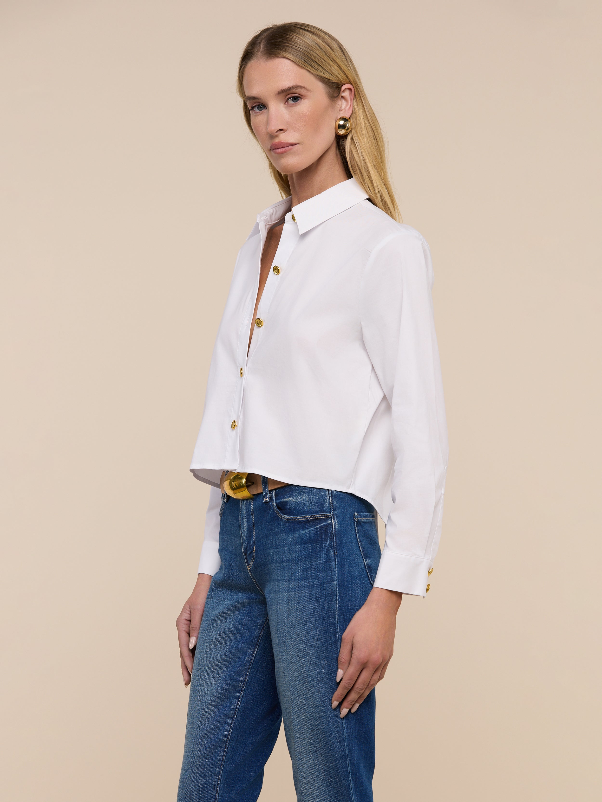 A woman with long blonde hair stands facing forward against a plain light background, wearing the Cosette Shirt in white poplin and blue jeans.