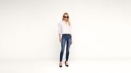 A woman wearing sunglasses, a white shirt, blue jeans, and black heels stands against a plain white background.