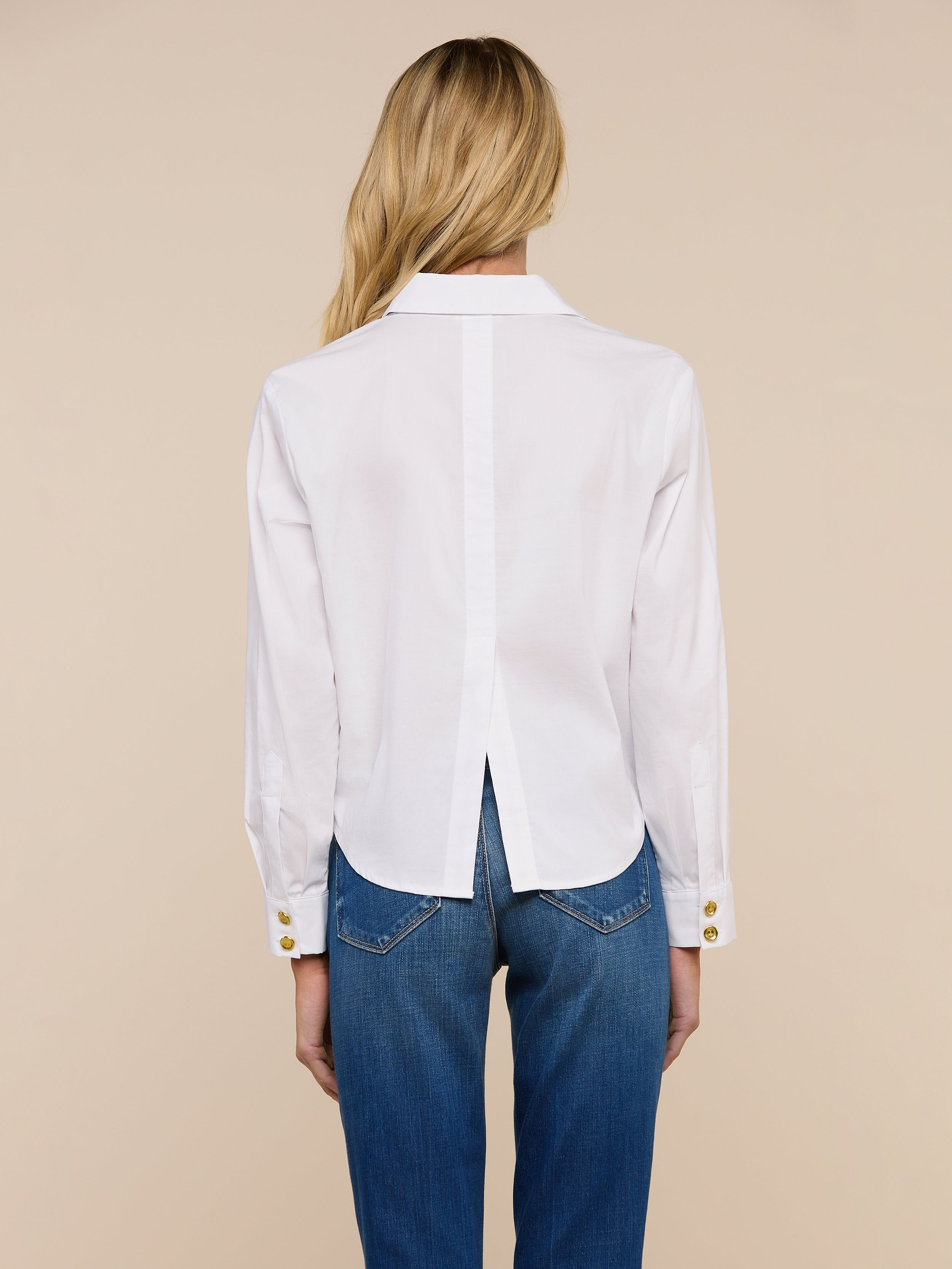 A person with long blonde hair, dressed in the Cosette Shirt and blue jeans, stands facing away from the camera against a plain light background.