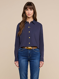 A woman with long brown hair stands facing forward against a plain light background, wearing a navy blue Cosette Shirt and blue jeans, her expression neutral.