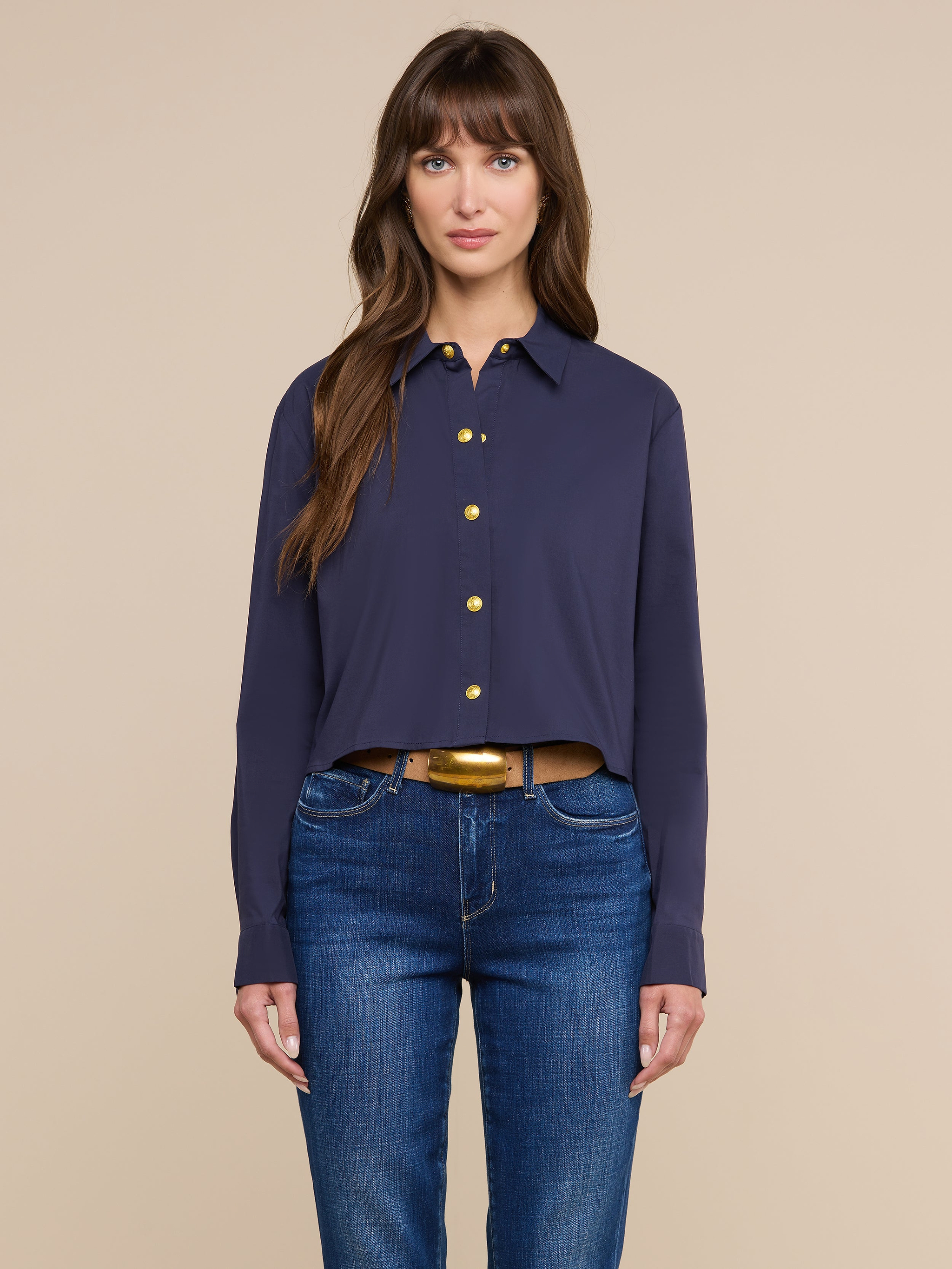 A woman with long brown hair stands facing forward against a plain light background, wearing a navy blue Cosette Shirt and blue jeans, her expression neutral.