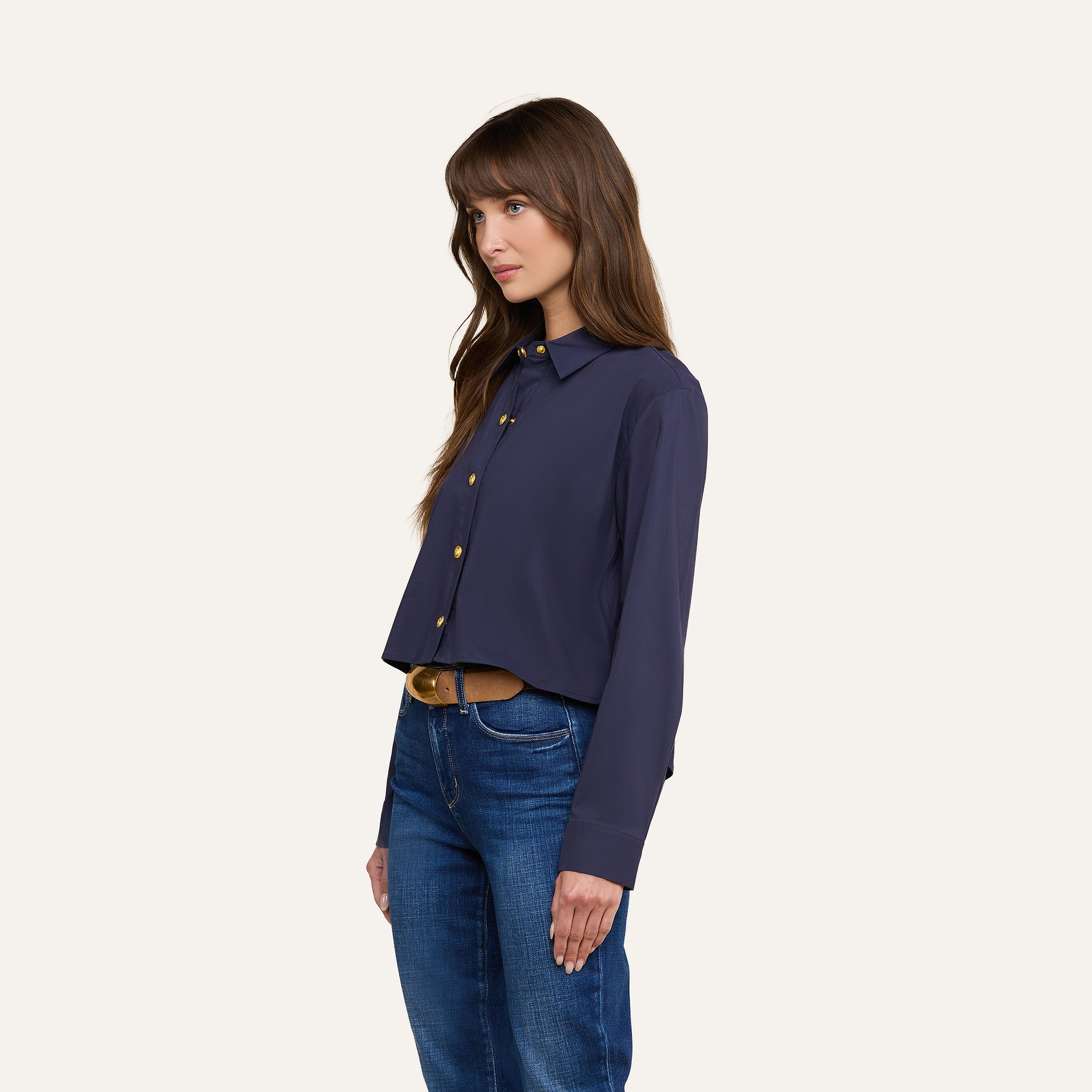 A woman with long brown hair wears the Cosette Shirt in navy, paired with blue jeans, standing against a plain light background and facing slightly left with a neutral expression.