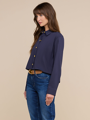 A woman with long brown hair wears the Cosette Shirt—navy blue, tailored, and made from organic cotton poplin—with blue jeans. She stands against a plain beige background, looking to the side.