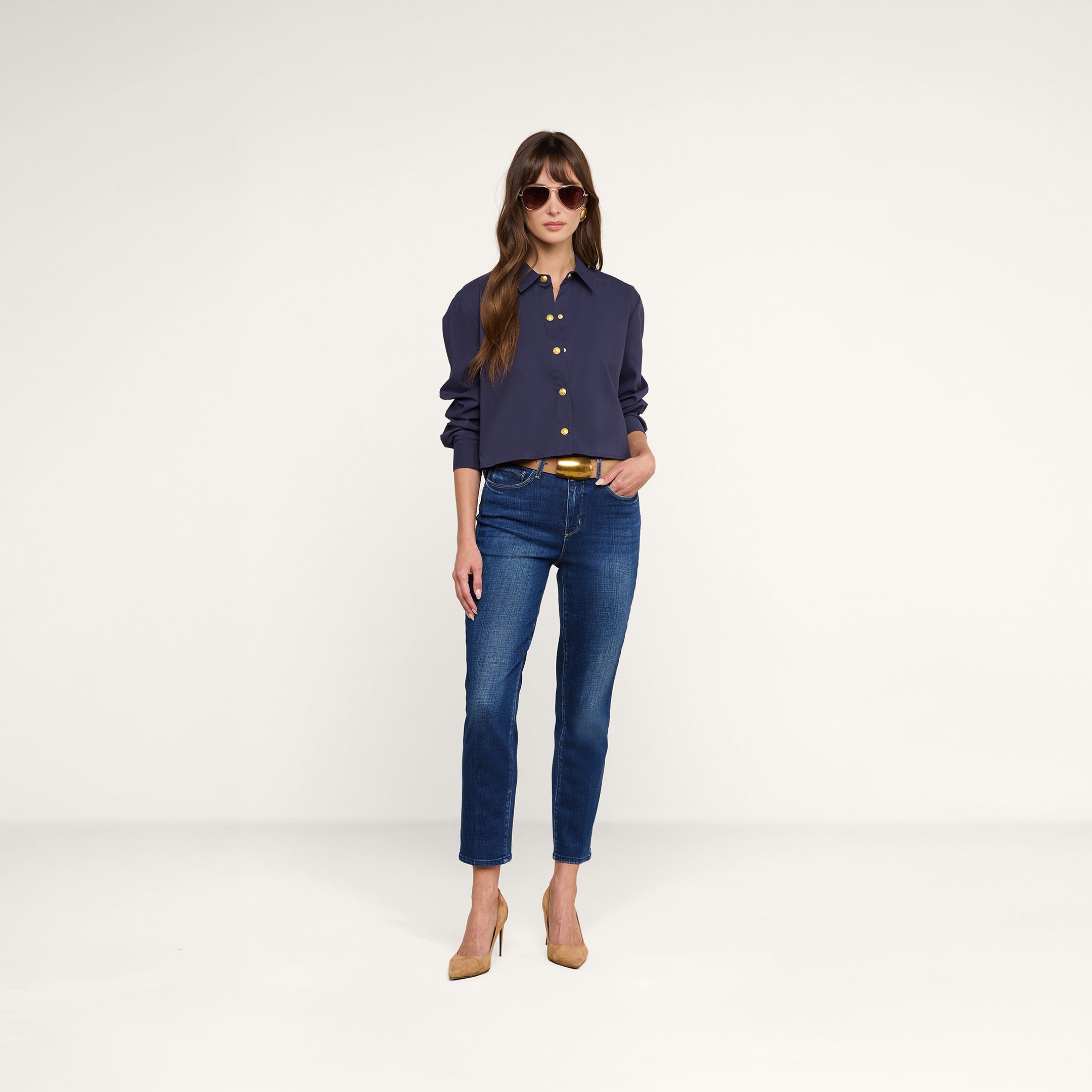 A woman stands against a plain white background, hands in pockets and looking forward, wearing sunglasses, blue jeans, beige heels, and the Cosette Shirt in navy.