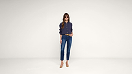A woman wearing sunglasses, a dark long-sleeve shirt, blue jeans, and tan heels stands against a plain white background with one hand in her pocket.
