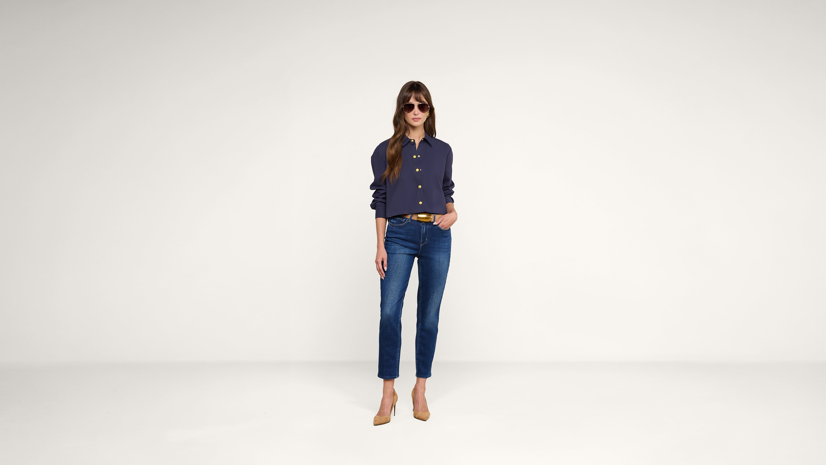 A woman wearing sunglasses, a dark long-sleeve shirt, blue jeans, and tan heels stands against a plain white background with one hand in her pocket.