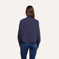 A woman with long brown hair is shown from the back, wearing the Cosette Shirt—a dark blue tailored shirt with gold buttons on the cuffs—paired with blue jeans, standing against a plain light background.