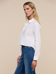 A woman with long blonde hair wears the Cosette Shirt and blue jeans, standing sideways against a plain beige background and looking slightly toward the camera.