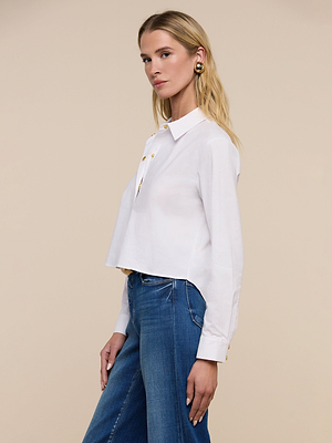 A woman with long blonde hair wears the Cosette Shirt and blue jeans, standing sideways against a plain beige background and looking slightly toward the camera.