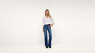 A person stands against a plain white background, wearing a white button-up shirt, blue flared jeans, and black shoes. They have long blonde hair and a neutral expression.