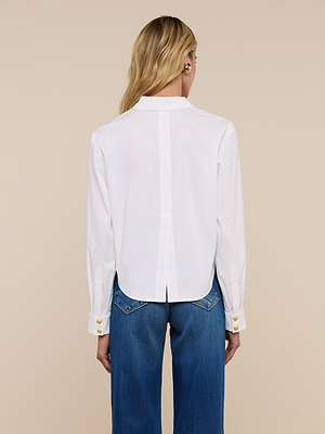 A person with long blonde hair, seen from behind against a beige background, is wearing the Cosette Shirt—an organic white cotton top with gold buttons—paired with blue jeans.