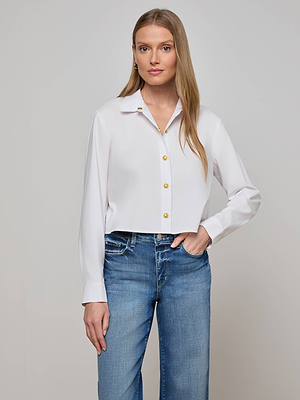 A woman with long blonde hair wears the L'AGENCE Cosette Shirt, a tailored white button-up with gold buttons, paired with blue jeans against a plain light gray background.