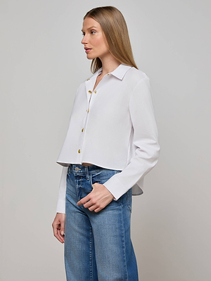 A woman with straight, light brown hair models the L'AGENCE Cosette Shirt—an organic cotton poplin button-up—paired with blue jeans, standing sideways against a plain light gray background.