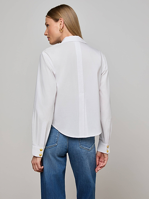 A person with straight blonde hair stands with their back to the camera against a light background, wearing L'AGENCE's Cosette Shirt—a tailored white poplin blouse with gold cuff buttons—paired with blue jeans.