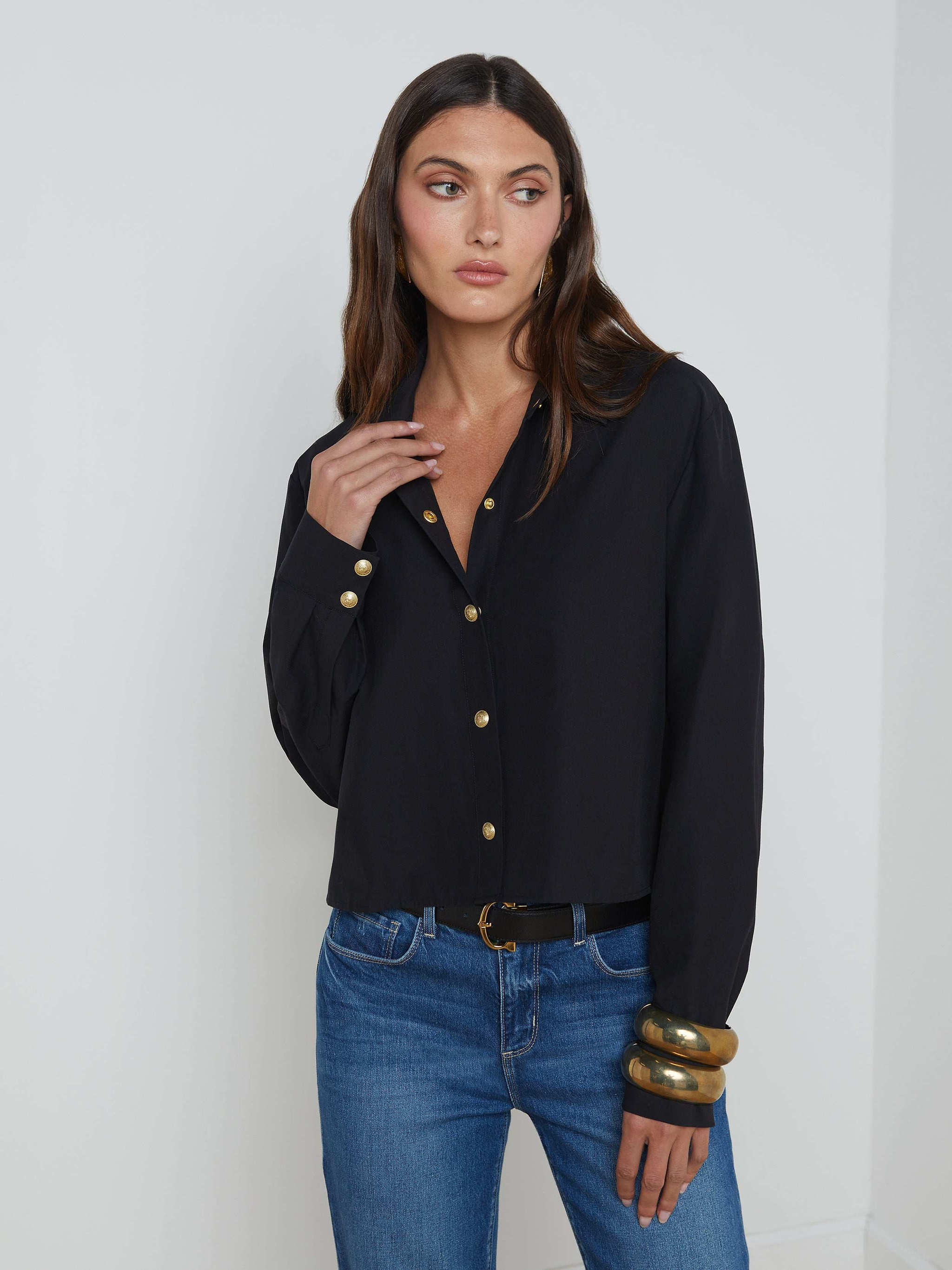Wearing the Cosette Shirt—a tailored black button-up—a woman with long brown hair stands against a plain background, looking to her left. She pairs the top with blue jeans and chunky gold bracelets.