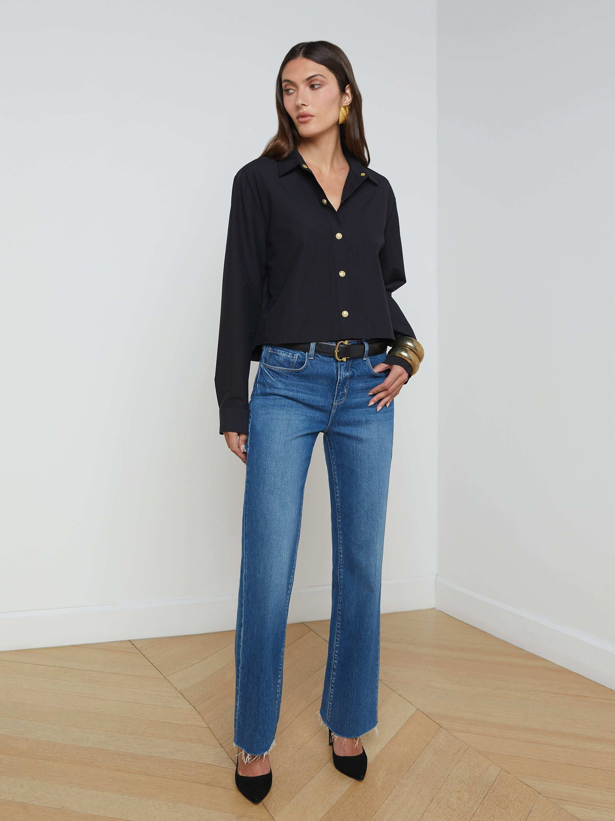 A woman stands against a plain white background wearing the Cosette Shirt, styled with blue jeans, a black belt, black heels, and large hoop earrings.