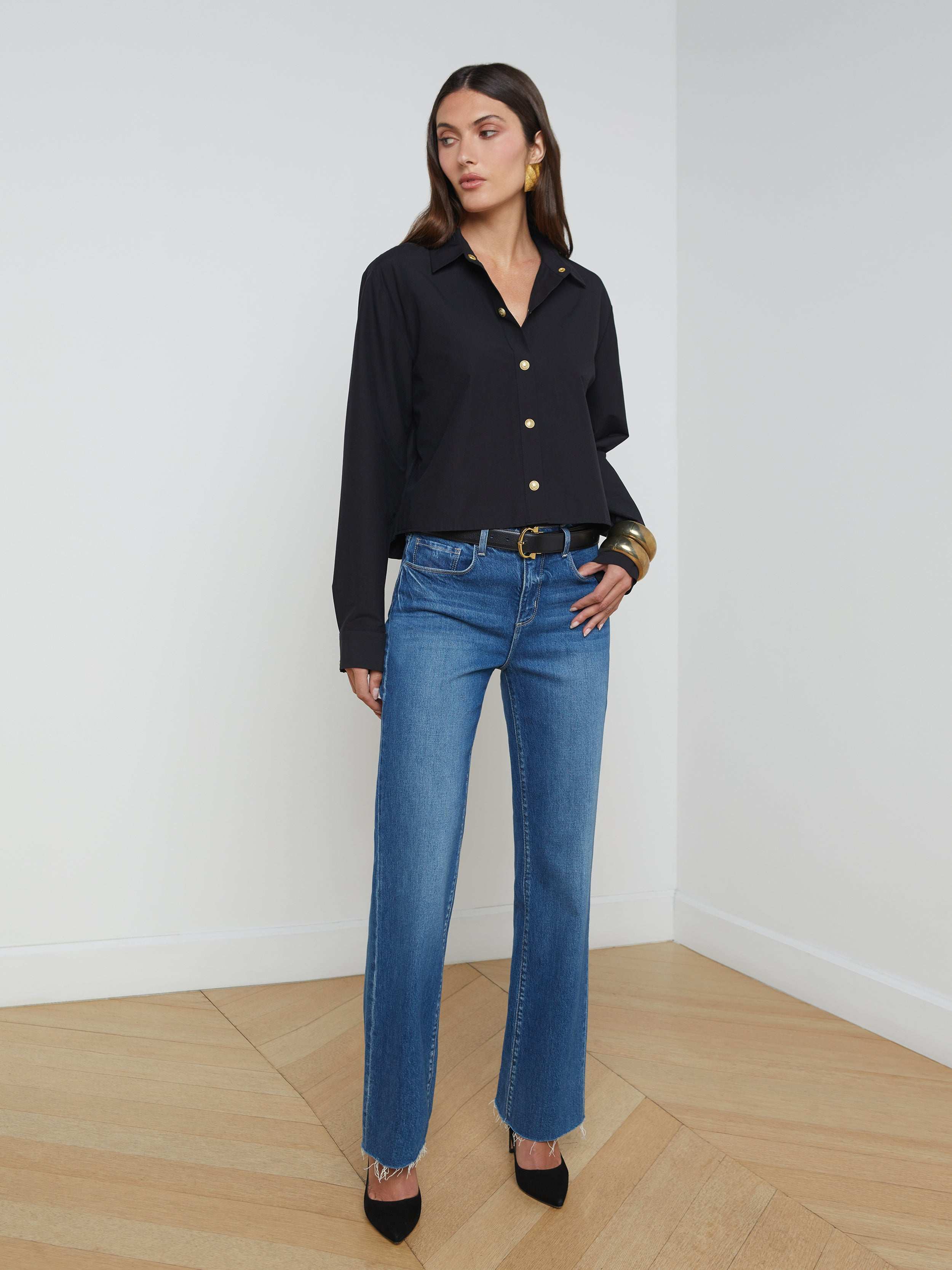 A woman stands against a plain white background wearing the Cosette Shirt, styled with blue jeans, a black belt, black heels, and large hoop earrings.