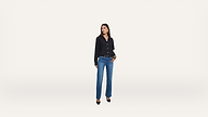 A woman stands against a plain white background, wearing a black button-up shirt, blue jeans, and black pointed-toe heels. She has dark hair and is looking slightly to the side.