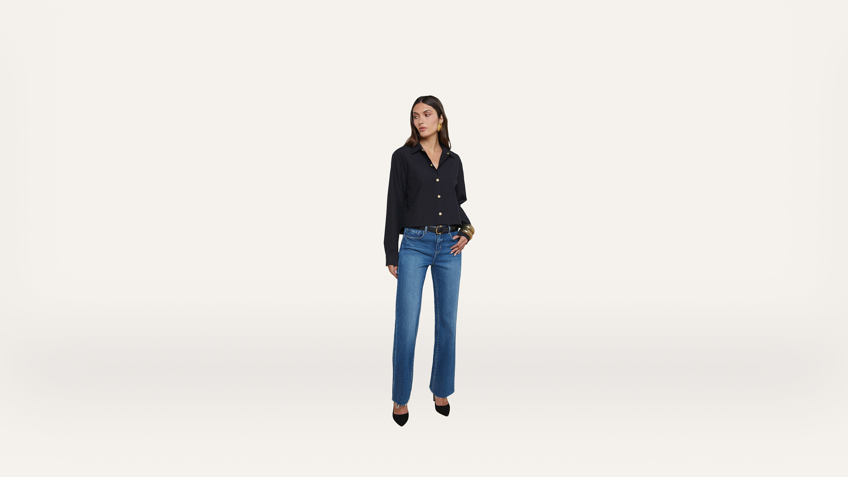 A woman stands against a plain white background, wearing a black button-up shirt, blue jeans, and black pointed-toe heels. She has dark hair and is looking slightly to the side.
