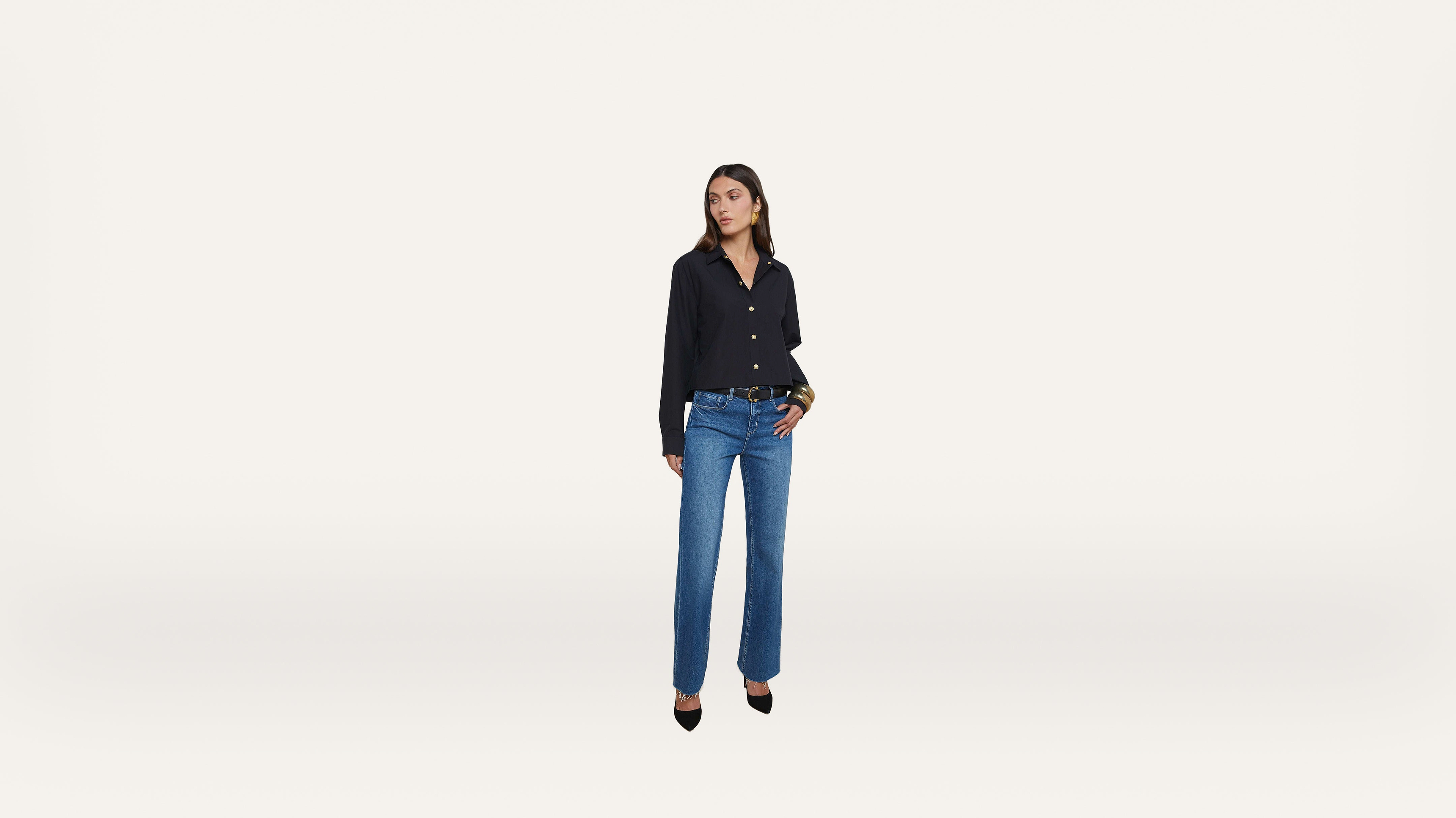 A woman stands against a plain white background, wearing a black button-up shirt, blue jeans, and black pointed-toe heels. She has dark hair and is looking slightly to the side.