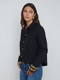 A woman with long brown hair wears the black Cosette Shirt, a stretch cotton-blend tailored top, paired with blue jeans and accented by large gold bracelets and gold earrings as she stands against a plain light background.