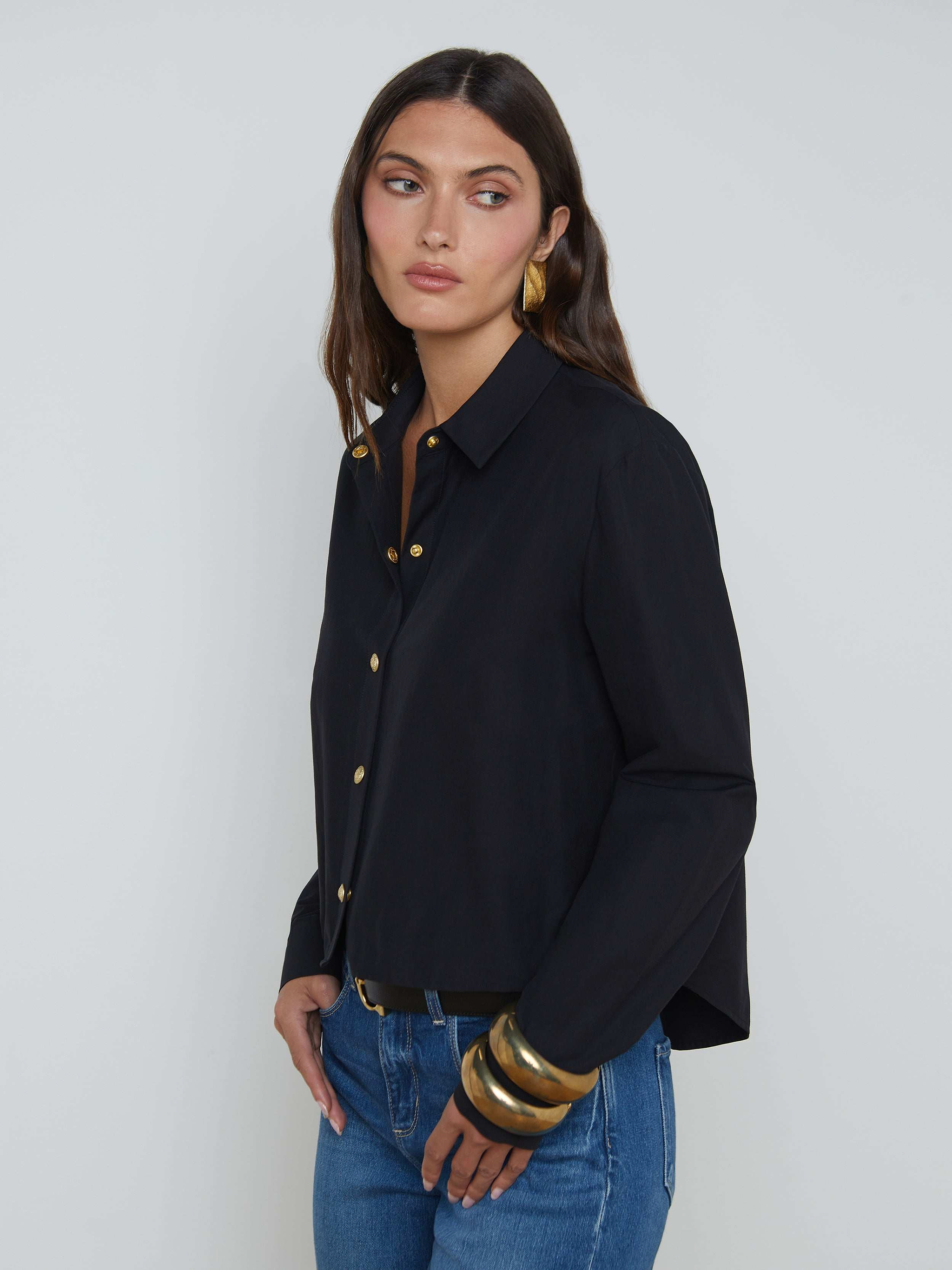 A woman with long brown hair wears the black Cosette Shirt, a stretch cotton-blend tailored top, paired with blue jeans and accented by large gold bracelets and gold earrings as she stands against a plain light background.