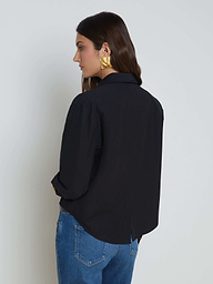 A woman with long brown hair wears the Cosette Shirt—a black stretch cotton-blend tailored design—paired with blue jeans and large gold earrings, standing facing away with her head turned slightly to the side against a plain light background.