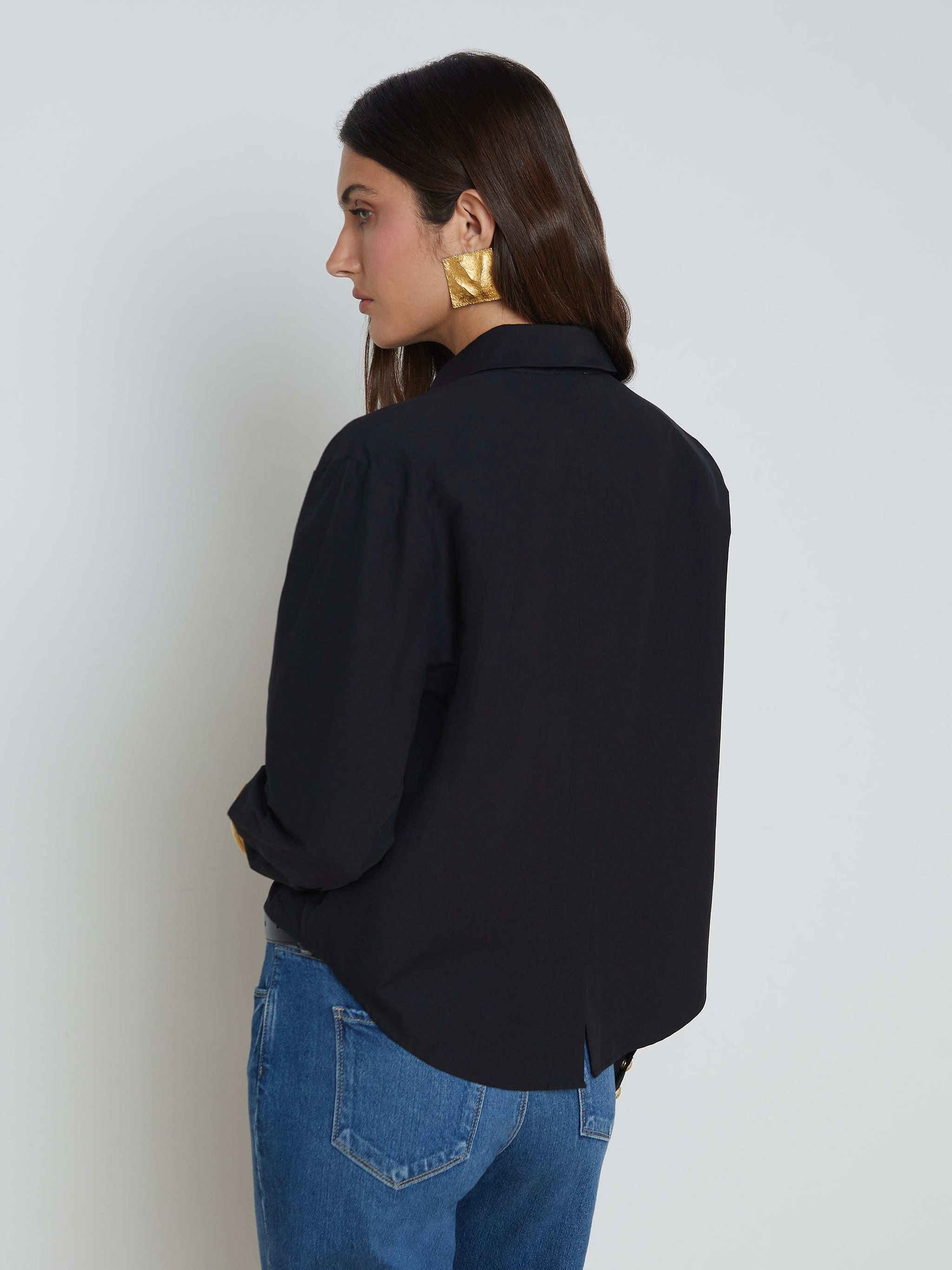 A woman with long brown hair wears the Cosette Shirt—a black stretch cotton-blend tailored design—paired with blue jeans and large gold earrings, standing facing away with her head turned slightly to the side against a plain light background.