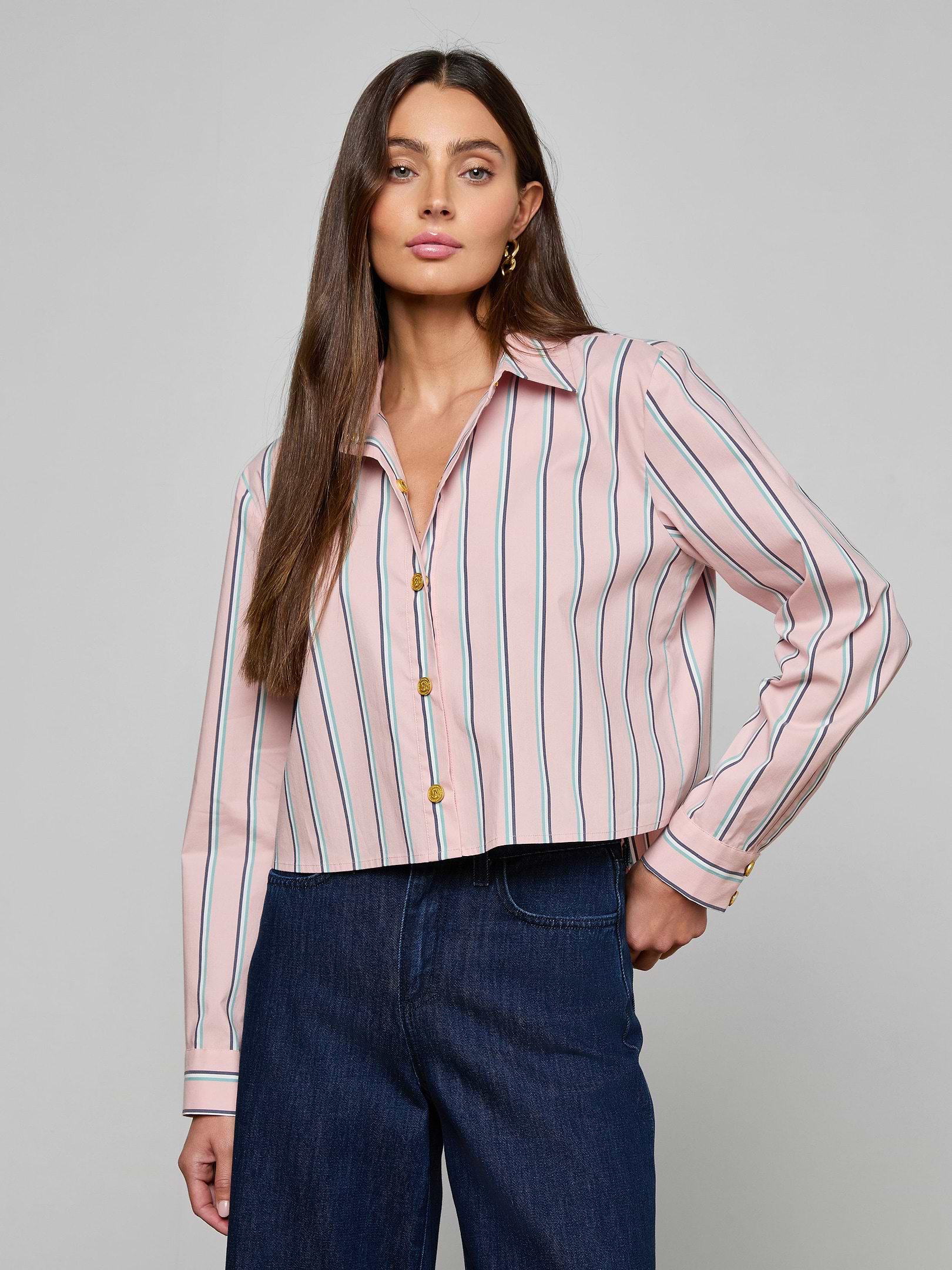 A woman with long brown hair is wearing the Cosette Shirt—a pink tailored shirt with vertical blue and red stripes, featuring a cropped high-low hem—paired with dark blue jeans and standing against a plain light background.