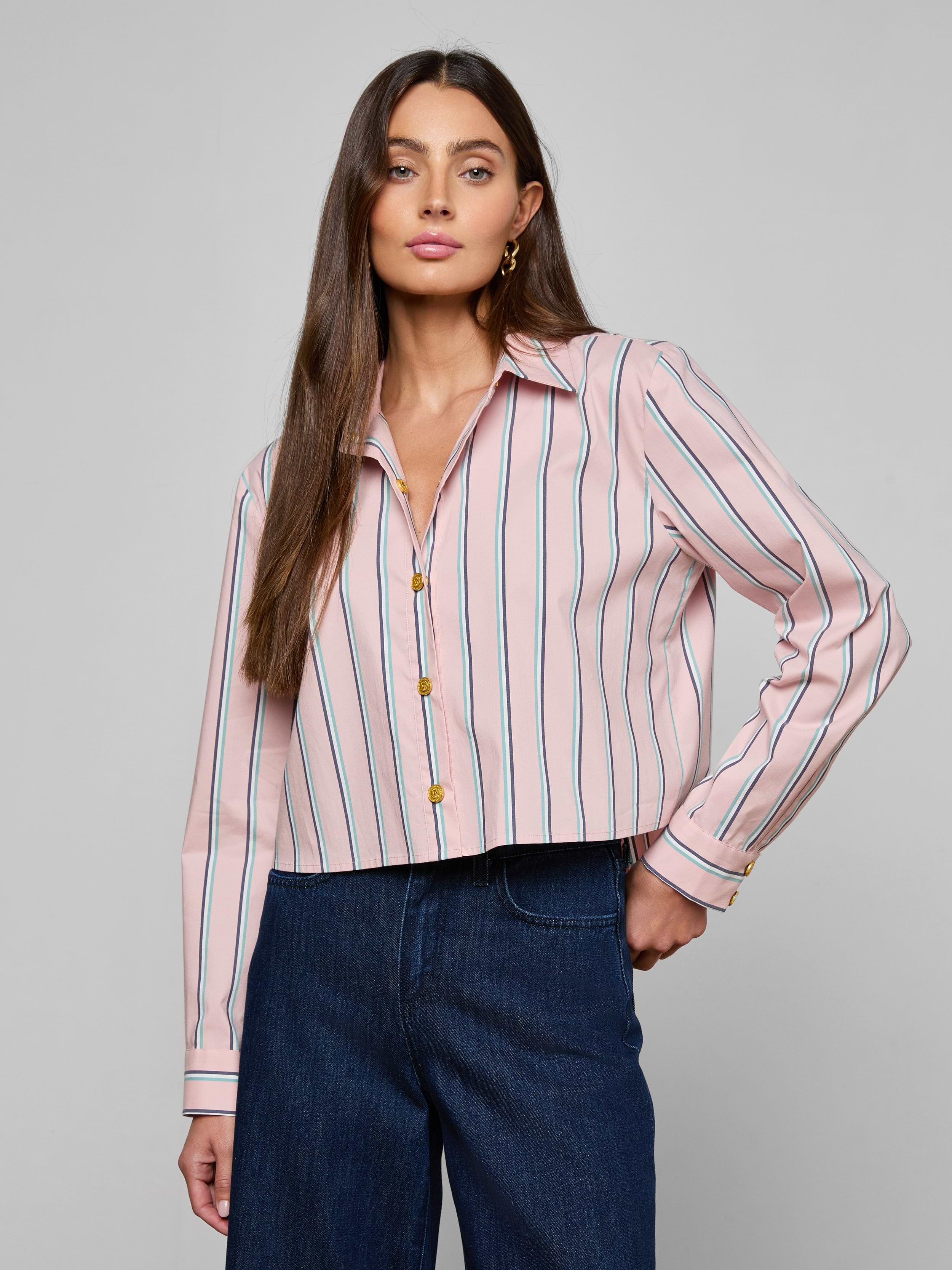 A woman with long brown hair is wearing the Cosette Shirt—a pink tailored shirt with vertical blue and red stripes, featuring a cropped high-low hem—paired with dark blue jeans and standing against a plain light background.