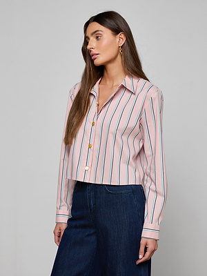 A woman with long brown hair wears the Cosette Shirt—an organic cotton-blend, pink tailored shirt with vertical stripes and long sleeves—paired with dark blue jeans, standing against a plain light gray background.