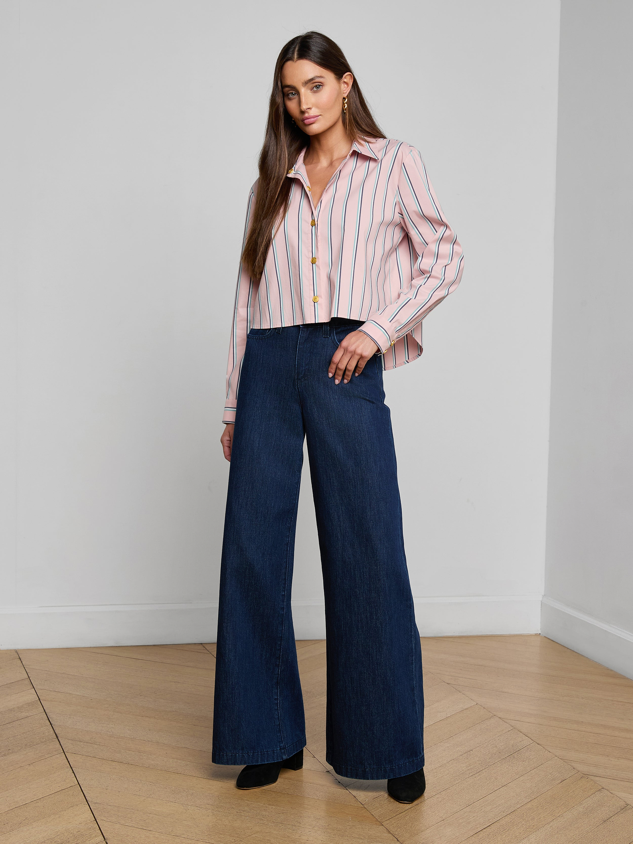 A woman stands against a plain white background wearing the Cosette Shirt—a tailored, striped button-up made from an organic cotton-blend—paired with high-waisted, wide-leg dark blue jeans and black shoes.