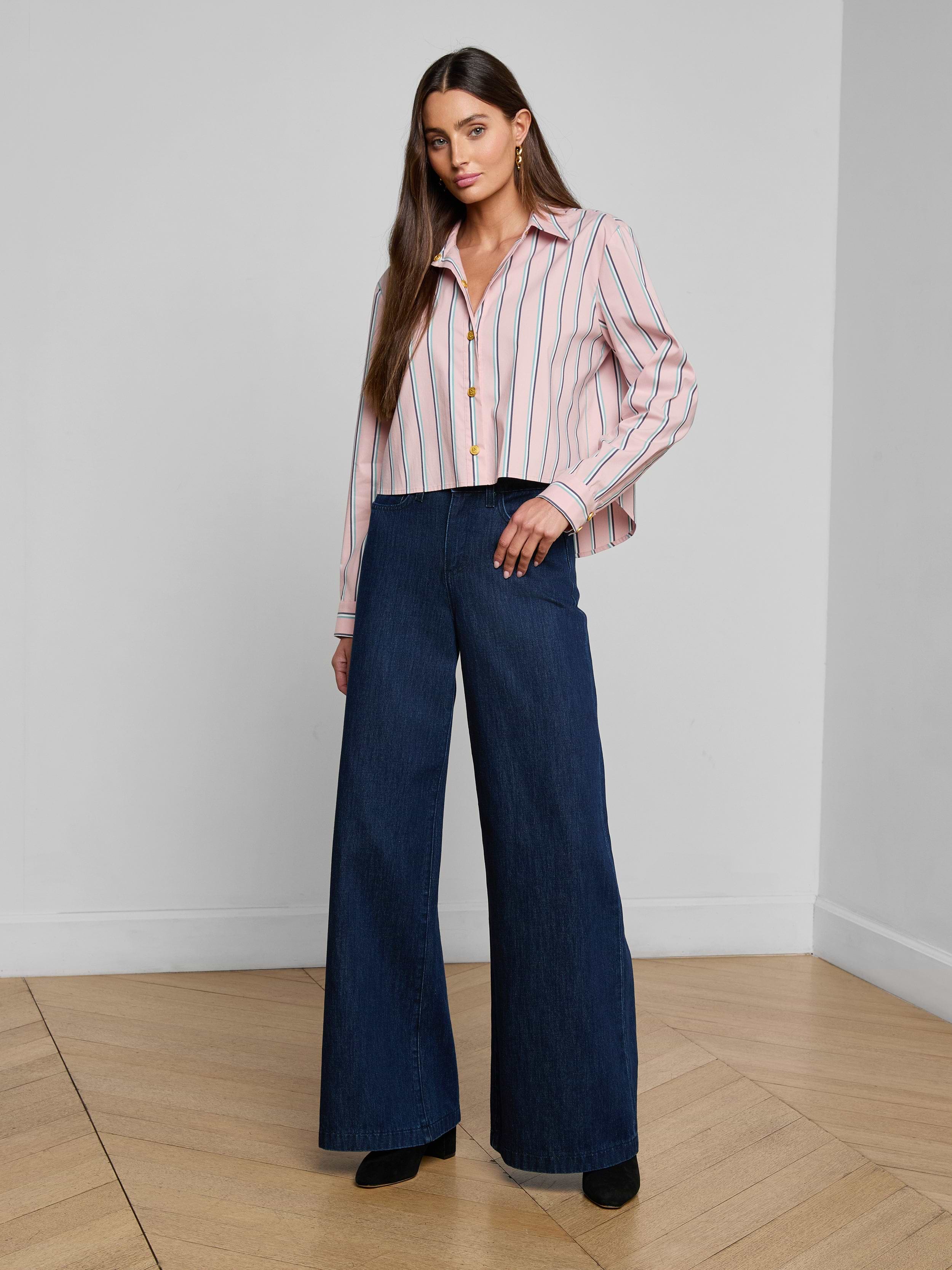 A woman stands against a plain white background wearing the Cosette Shirt—a tailored, striped button-up made from an organic cotton-blend—paired with high-waisted, wide-leg dark blue jeans and black shoes.