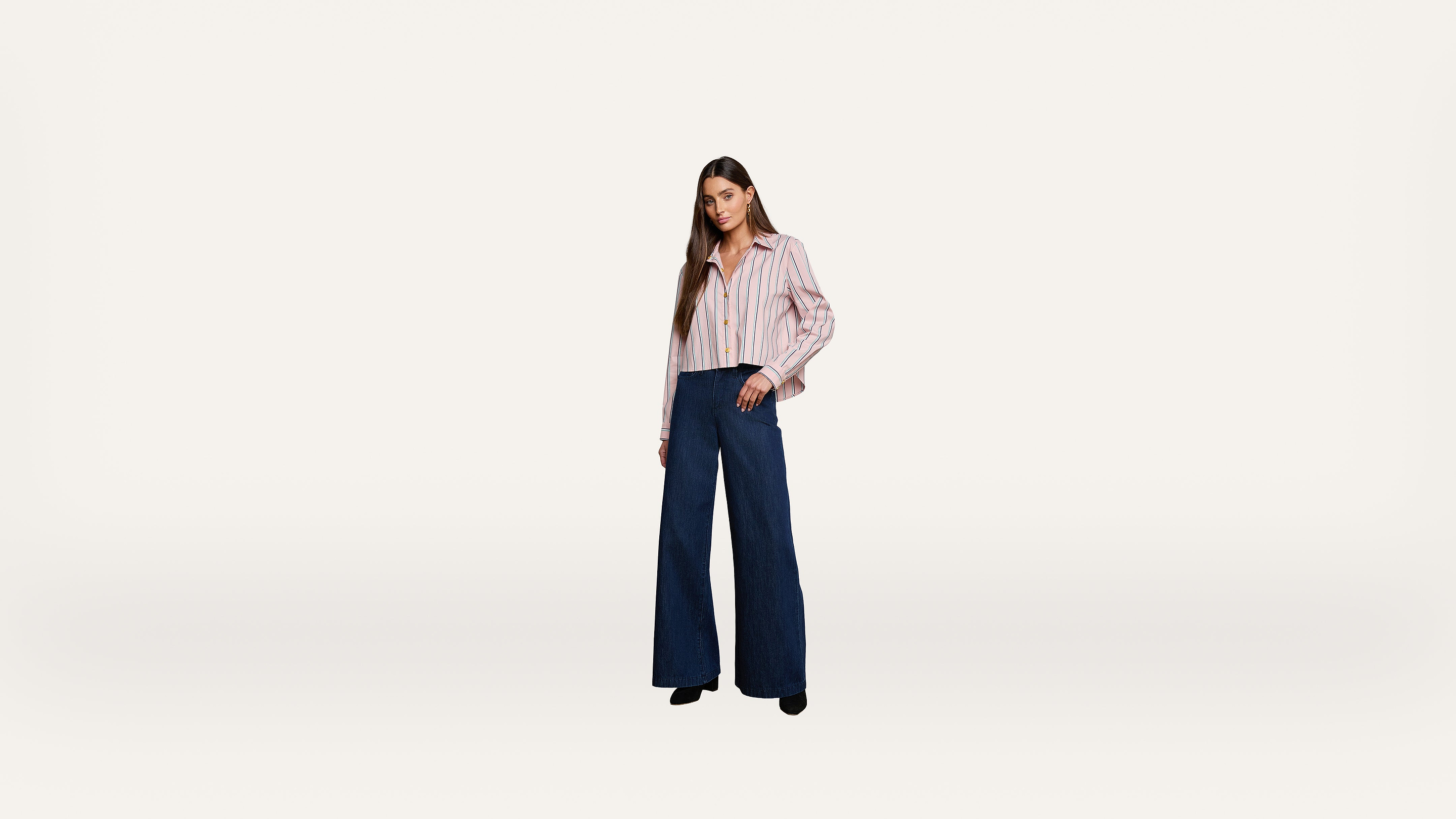 A woman stands against a plain white background, wearing a striped button-up shirt and high-waisted wide-leg blue pants with black shoes.