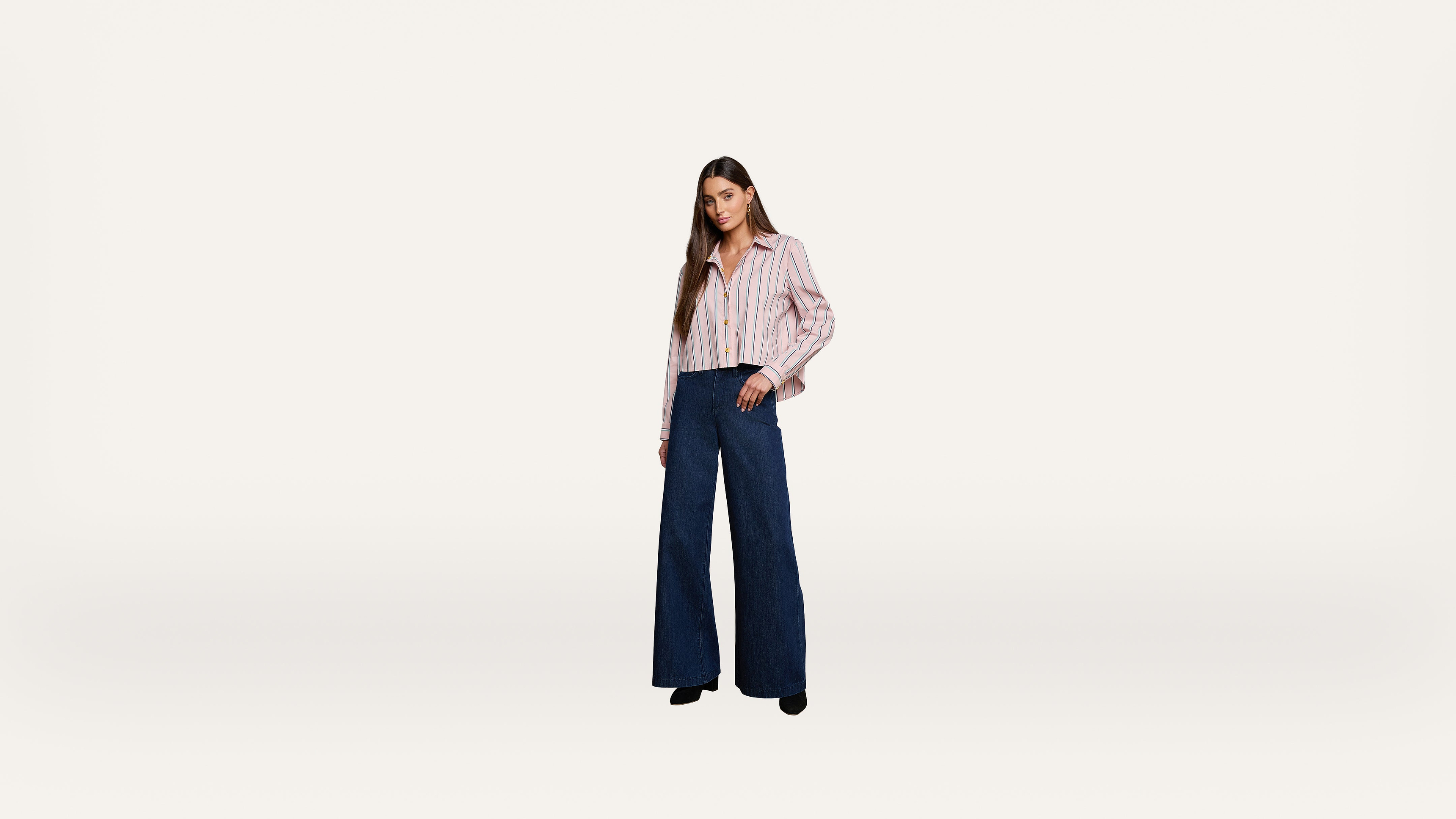 A woman stands against a plain white background, wearing a striped button-up shirt and high-waisted wide-leg blue pants with black shoes.