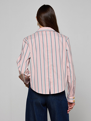 A woman with long brown hair faces away, modeling the Cosette Shirt—a tailored light pink top with blue and white vertical stripes, long sleeves, and a cropped high-low hem—paired with dark blue wide-leg jeans against a plain light background.