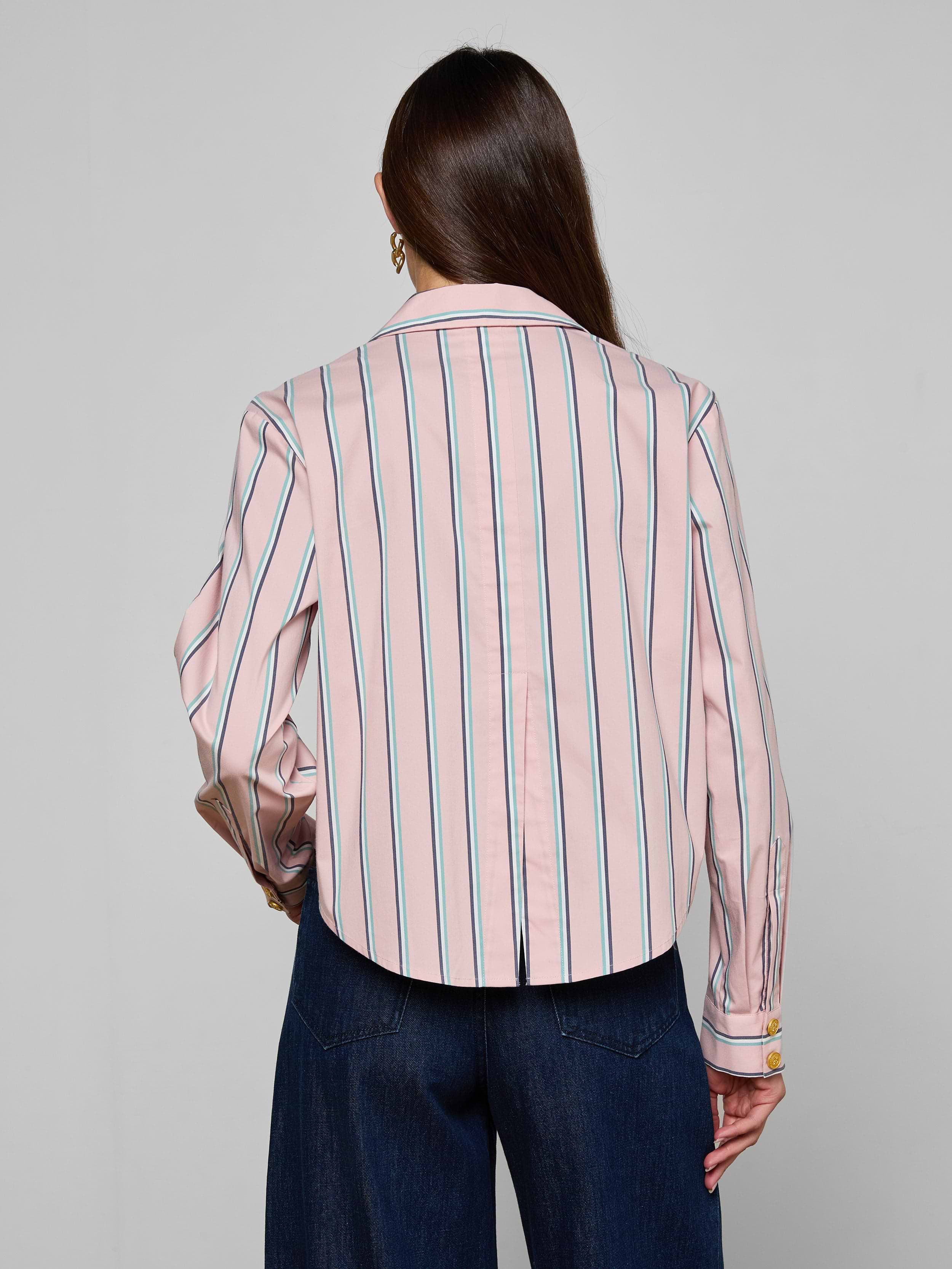 A woman with long brown hair is seen from behind, wearing the Cosette Shirt—a tailored pink, long-sleeve, collared shirt featuring vertical blue and white stripes and a cropped high-low hem—paired with dark blue wide-leg pants against a plain light background.