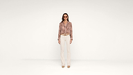 A woman stands facing forward wearing sunglasses, a patterned long-sleeve shirt, beige pants, a belt, and heels, against a plain white background.