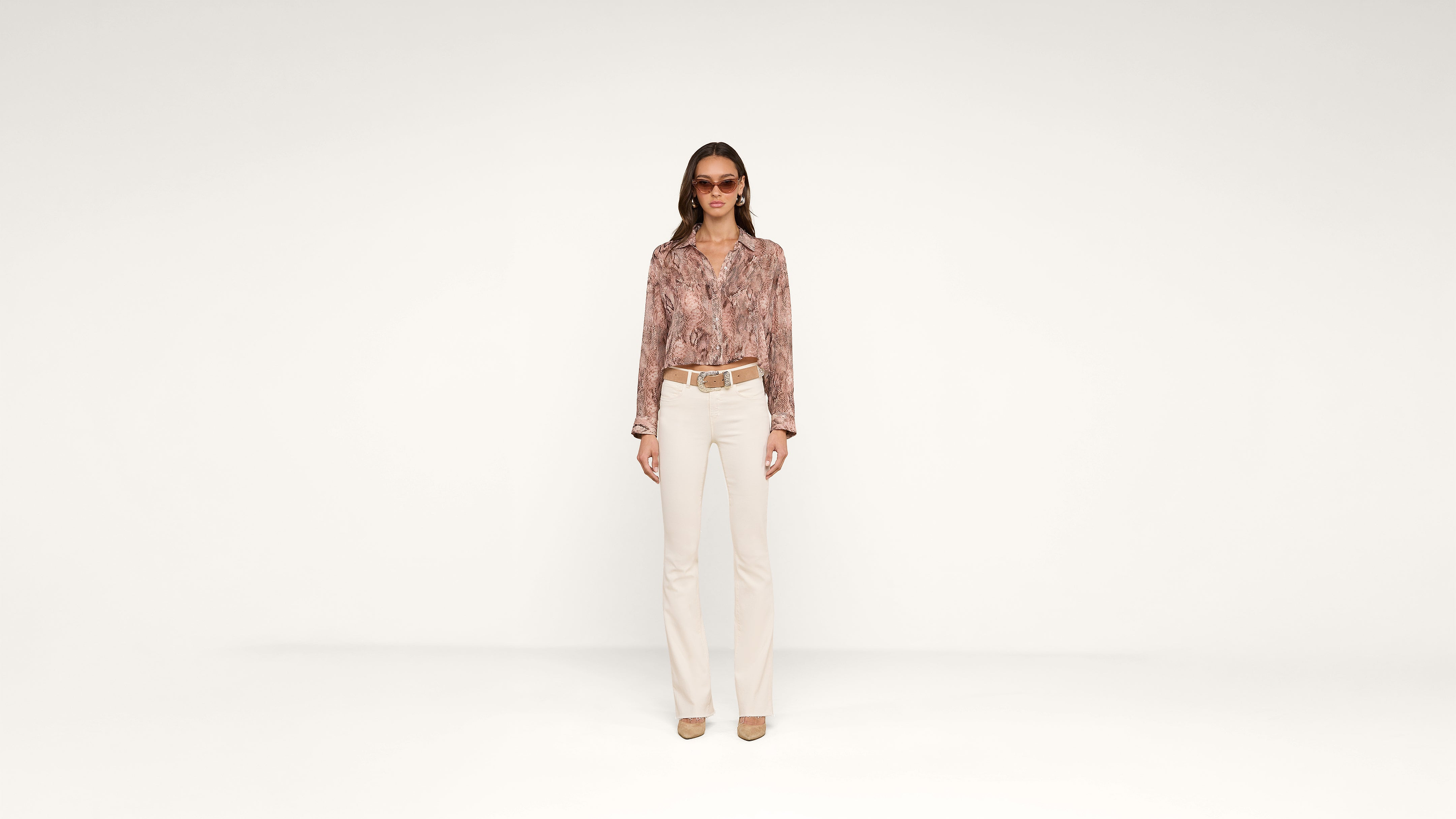 A woman stands facing forward wearing sunglasses, a patterned long-sleeve shirt, beige pants, a belt, and heels, against a plain white background.